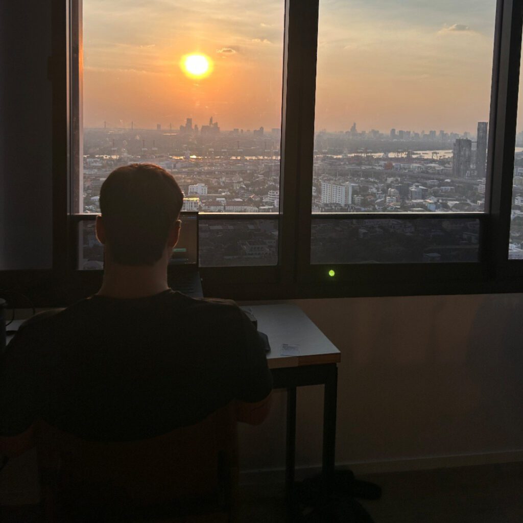 Digital nomad working from high-rise apartment in Bangkok, Thailand, overlooking city skyline at sunset — remote work with a view