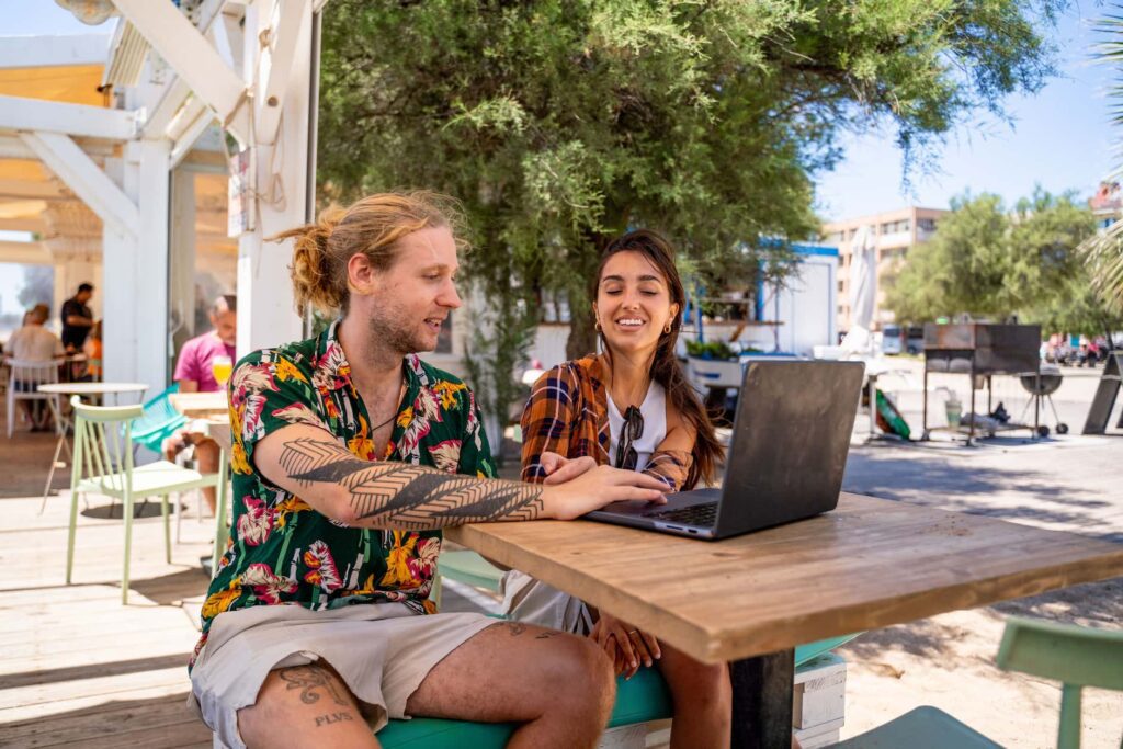 Digital nomads collaborating on a laptop at a beach café, showcasing the social and professional lifestyle of performance marketers abroad