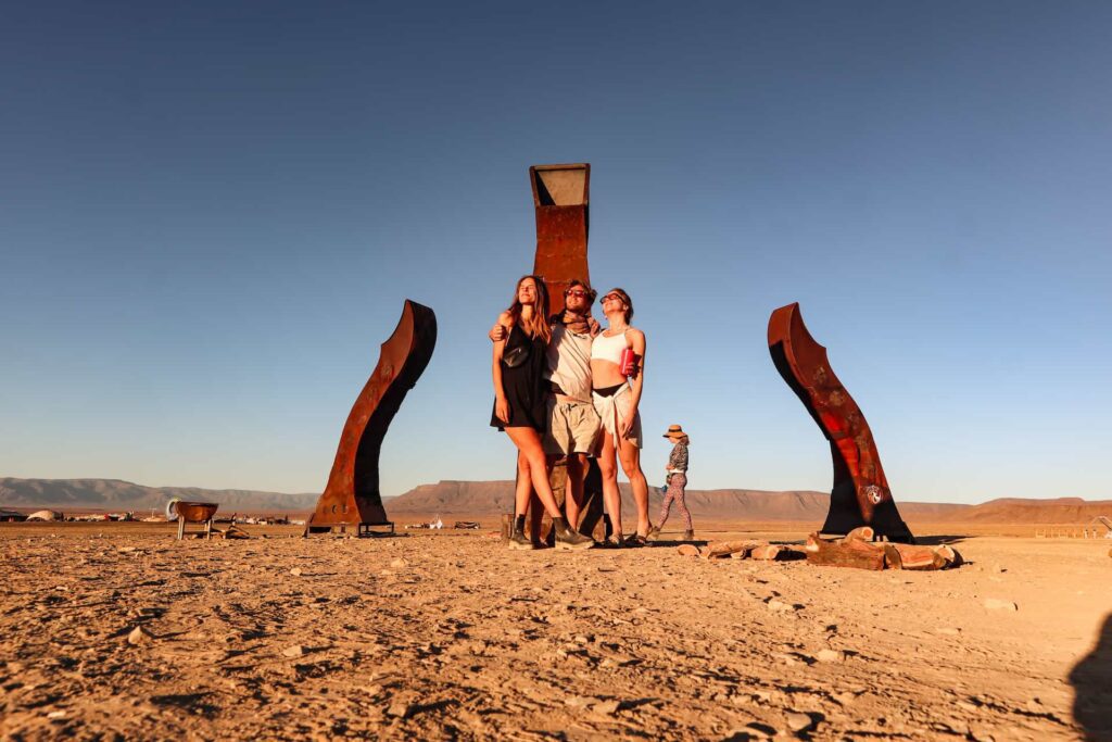 Friends posing at a large rusted steel art sculpture during golden hour at AfrikaBurn — blending creativity and connection in the Tankwa desert